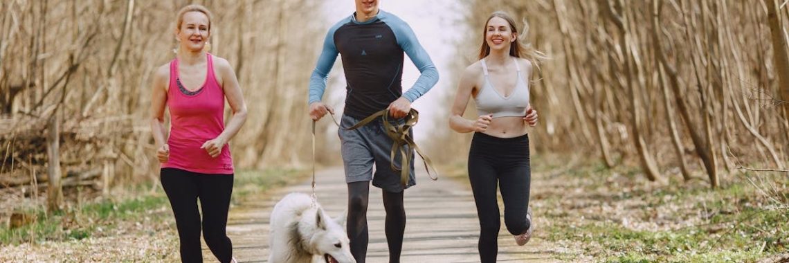 Three adults jogging with a dog on a forest path on a bright spring day, promoting healthy living.